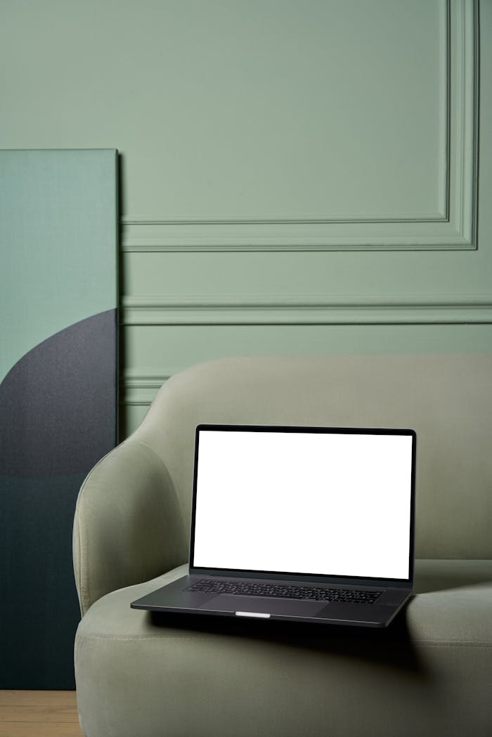 Laptop with white screen placed on soft sofa with big painting on floor near green uneven wall in bright apartment