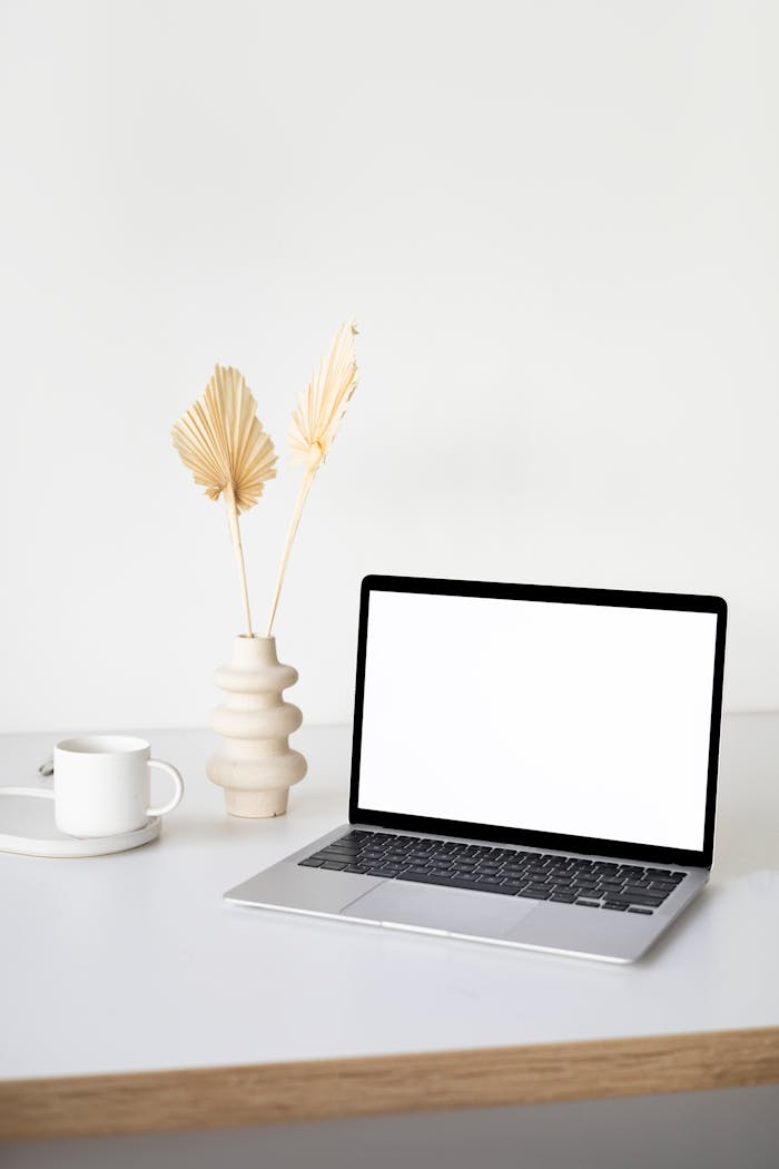 A clean and simple workspace setup with a laptop, ceramic vase, and coffee cup on a wooden table.